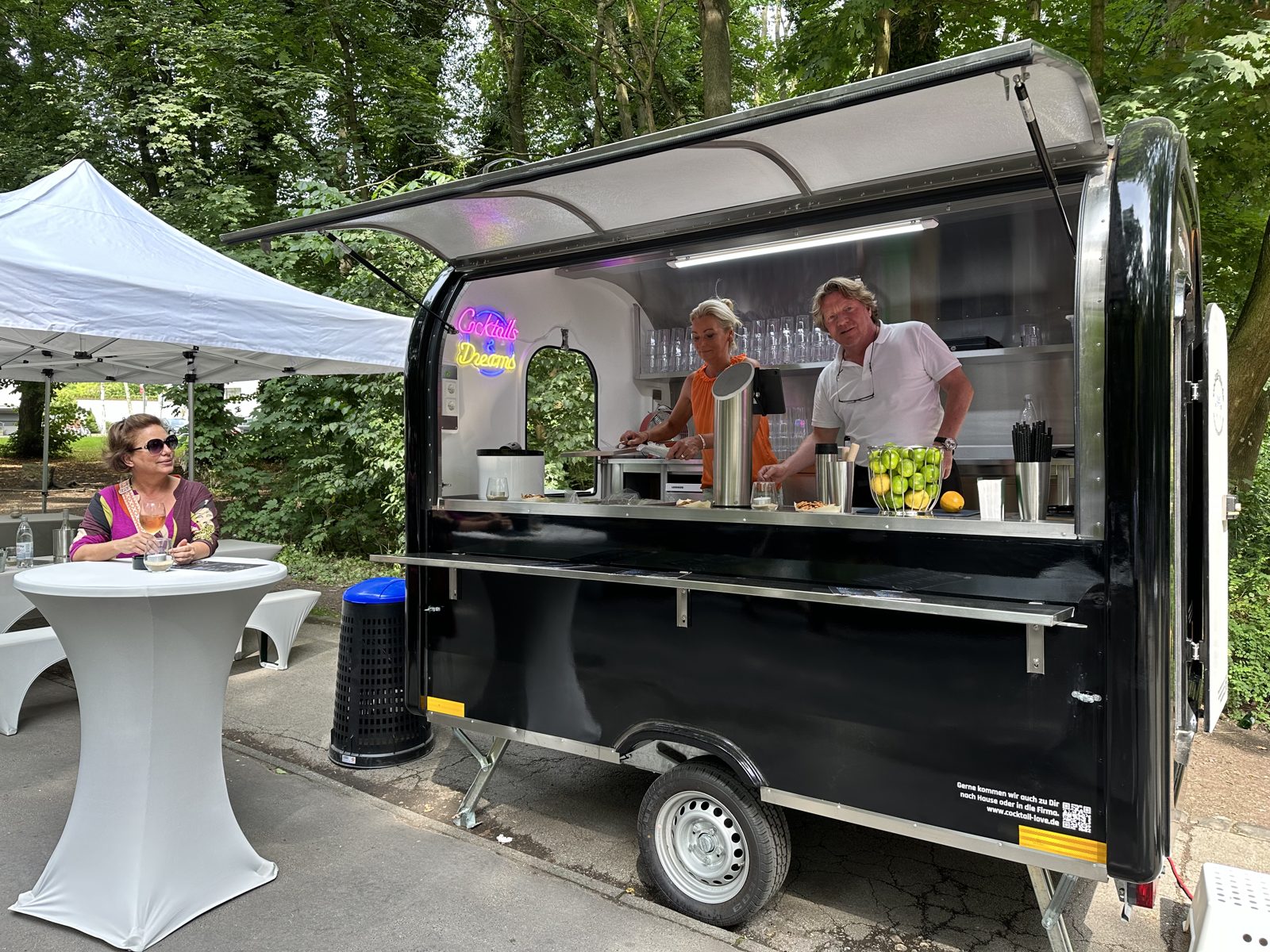 Der Cocktail-Trailer aufgebaut auf einer Veranstaltung, mit geöffneter Klappe, Tresen und Pavillon daneben.